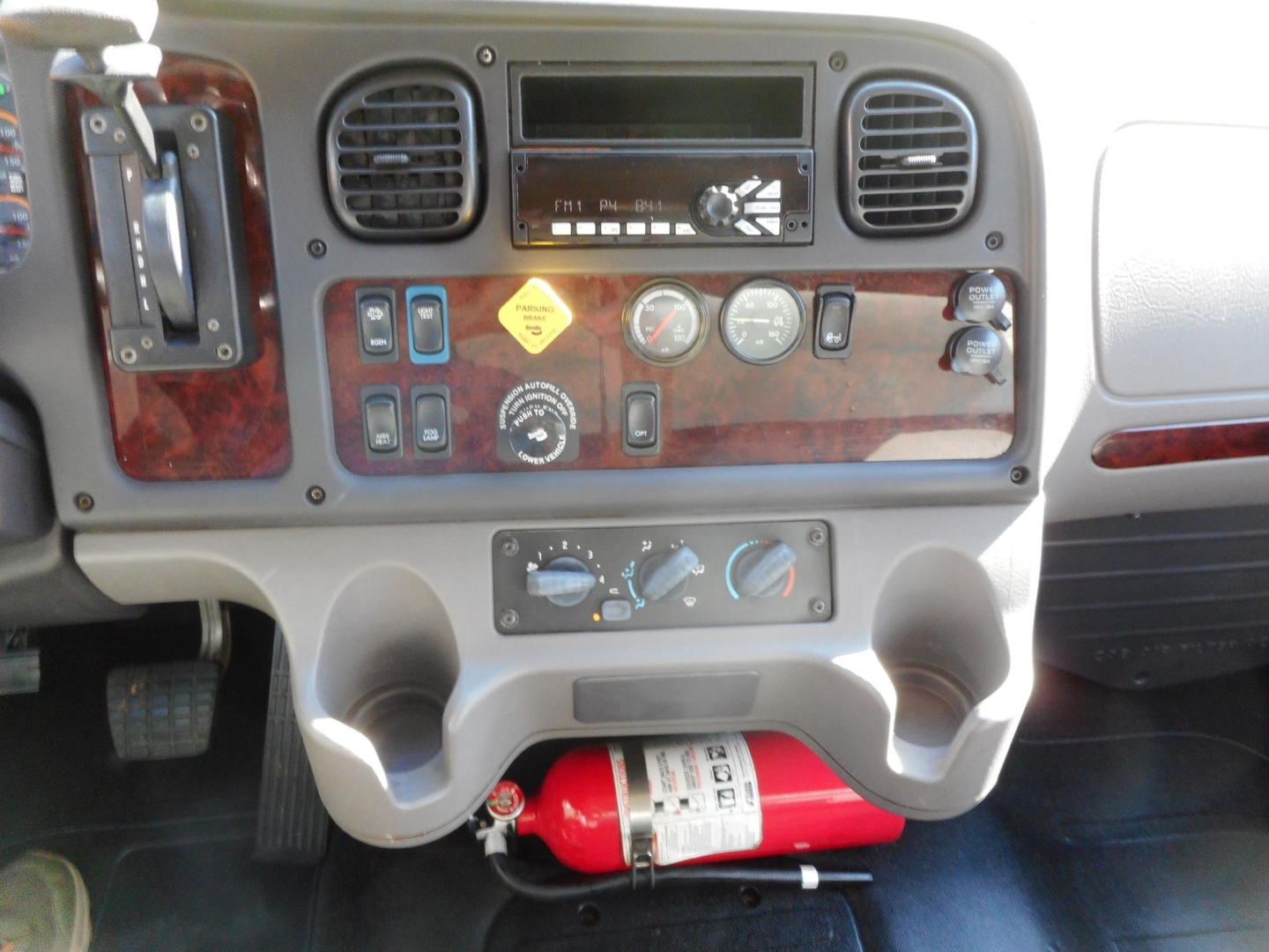 Dashboard view of a 2019 Freightliner M2 106 with woodgrain panel, gauges, FM radio, control switches, and a red fire extinguisher mounted under the console.