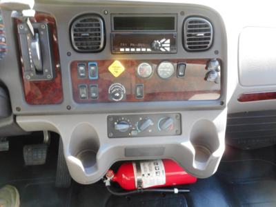 Dashboard view of a 2019 Freightliner M2 106 with woodgrain panel, gauges, FM radio, control switches, and a red fire extinguisher mounted under the console.