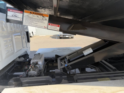Chevrolet 6500 HD truck chassis with hydraulic components and warning stickers under the dump bed, visible VIN and chassis wiring in foreground.