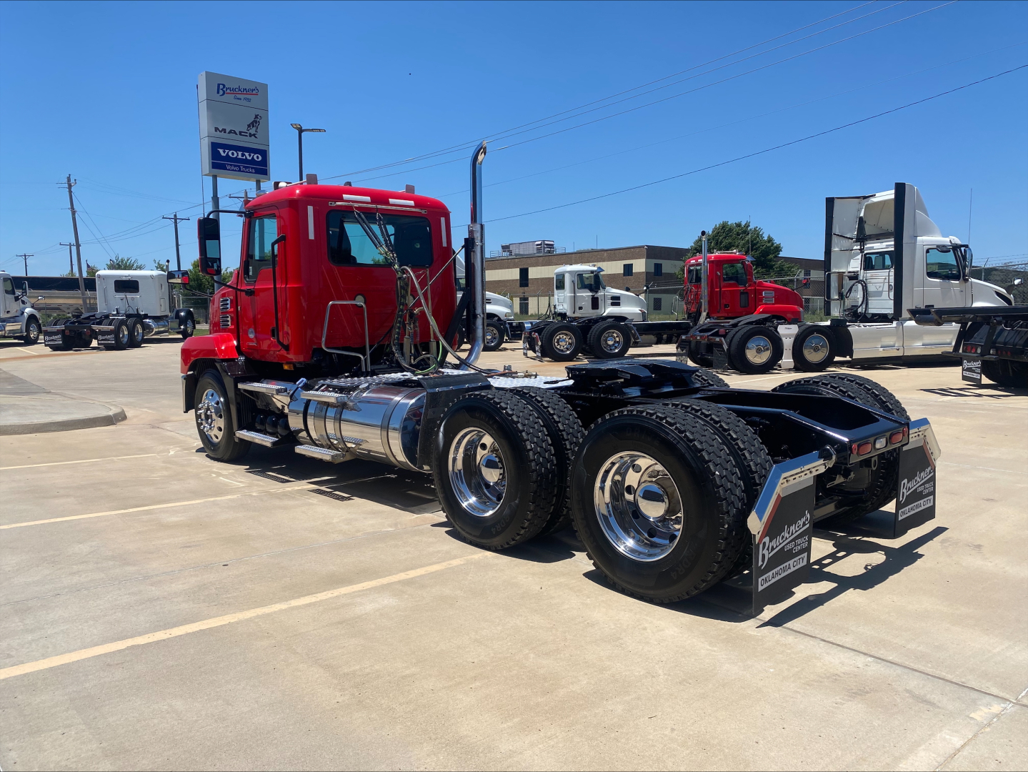 Truck chassis in a dealership lot with red Mack AN64T cab and dual rear axles, fuel tanks, and chrome wheels visible.