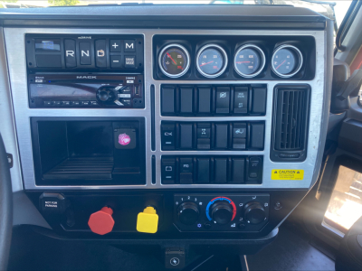 Dashboard of a Mack truck showing gear selector R N D plus M, six round gauges, various switches, a CD player/ radio, hazard switch, two colored knobs (red/ yellow) and climate controls.