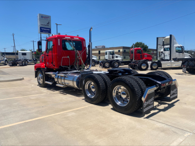 Truck chassis in a dealership lot with red Mack AN64T cab and dual rear axles, fuel tanks, and chrome wheels visible.