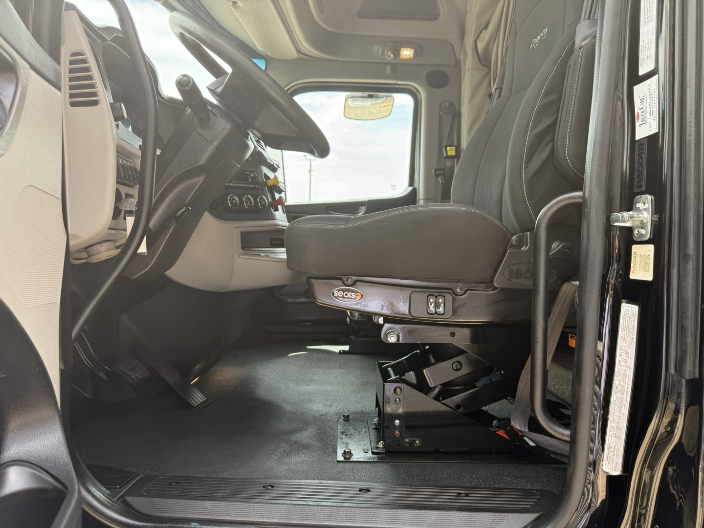 Truck cab interior of a 2022 Peterbilt 579 conventional sleeper, driver seat with suspension base, dashboard, steering wheel, and entry door visible.