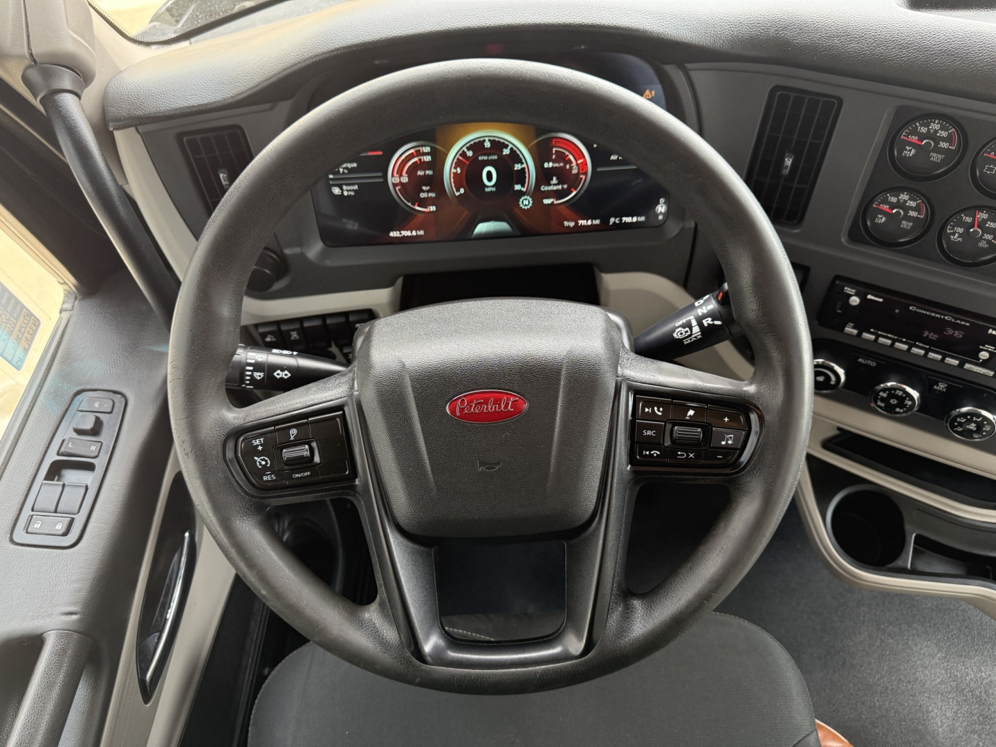 Peterbilt 579 cockpit view showing steering wheel with Peterbilt logo, digital instrument cluster, and dashboard gauges in front of a gray interior.