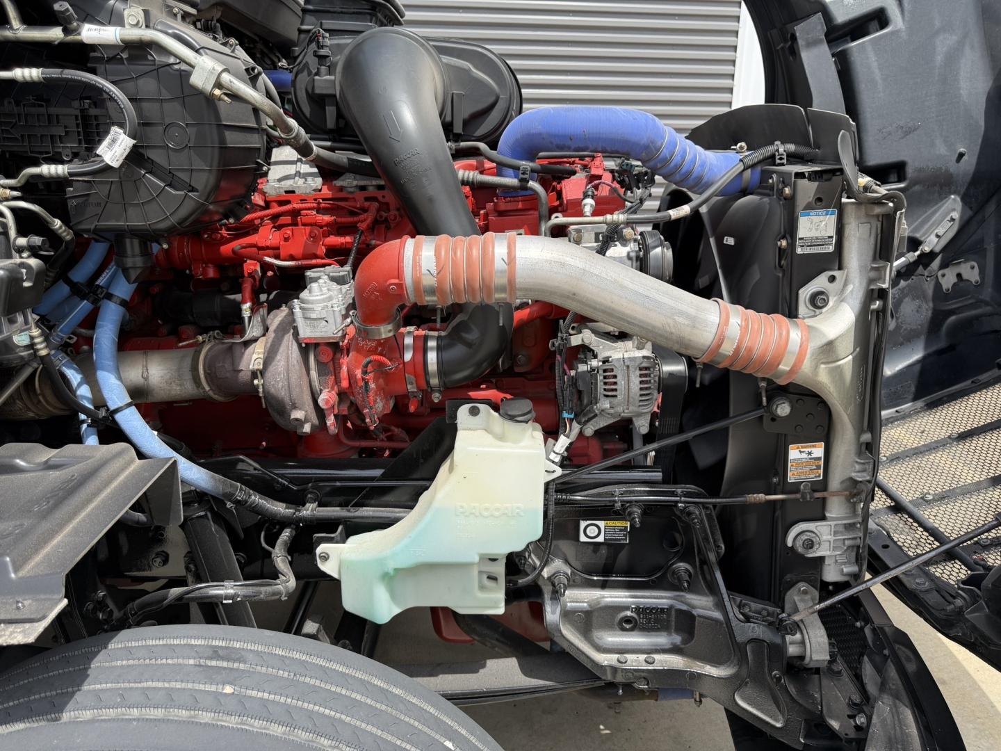 Peterbilt truck engine bay showing red engine, orange hoses, blue hoses, radiator, coolant reservoir, and various metal components.