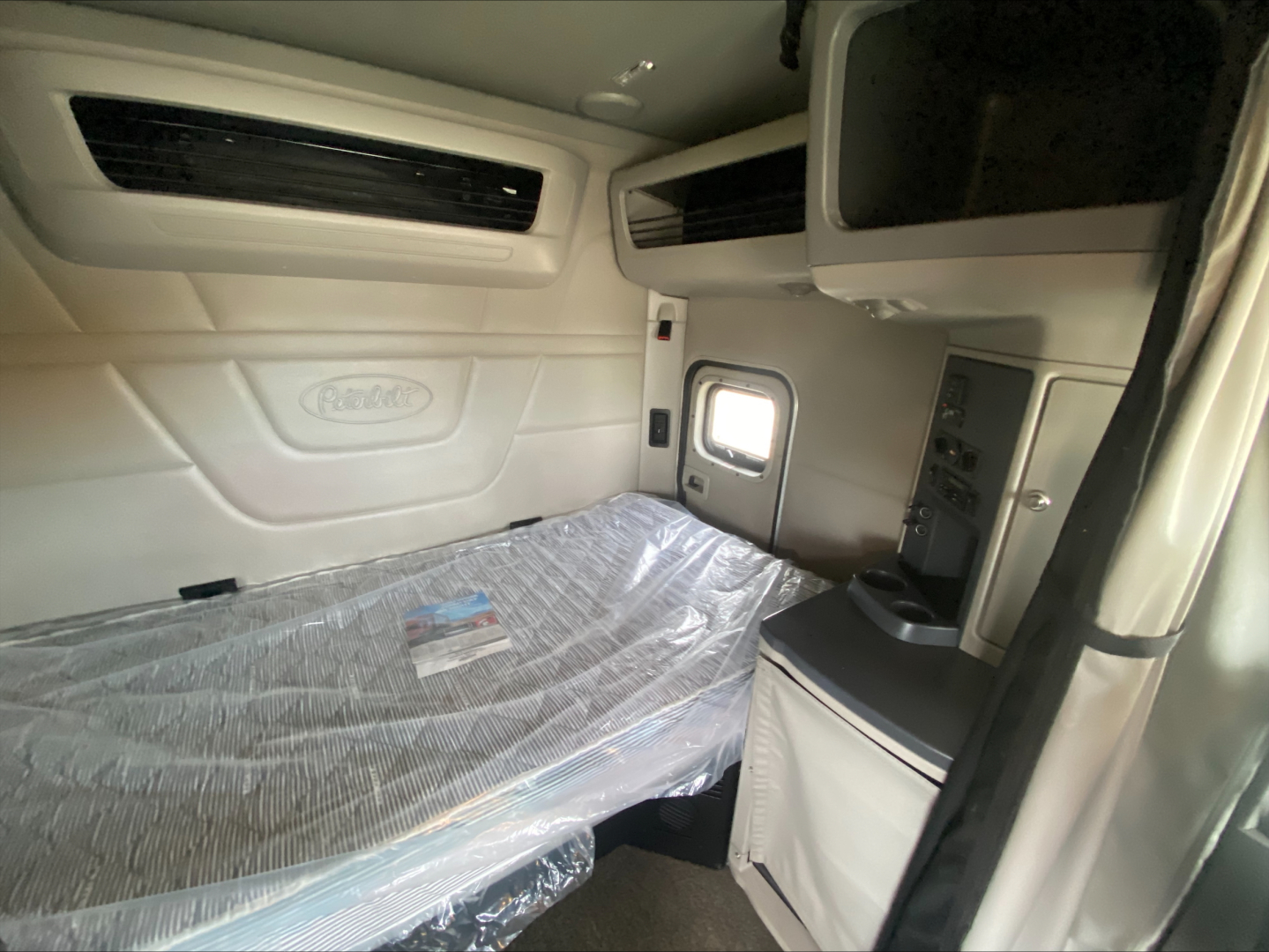 Peterbilt truck sleeper cab interior with bed and control panel; plastic-wrapped mattress visible.