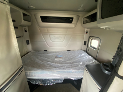 Truck cab interior view showing sleeping berth with mattress, light-colored panels, storage compartments, and small window.