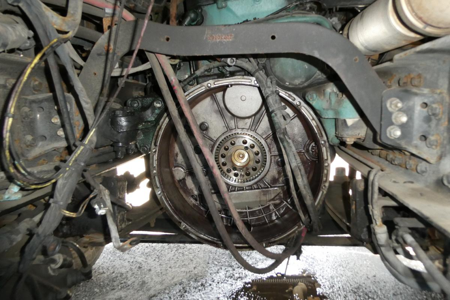 View under a Volvo VNM42T front axle showing drivetrain components and a circular flywheel housing, belts and hoses visible.