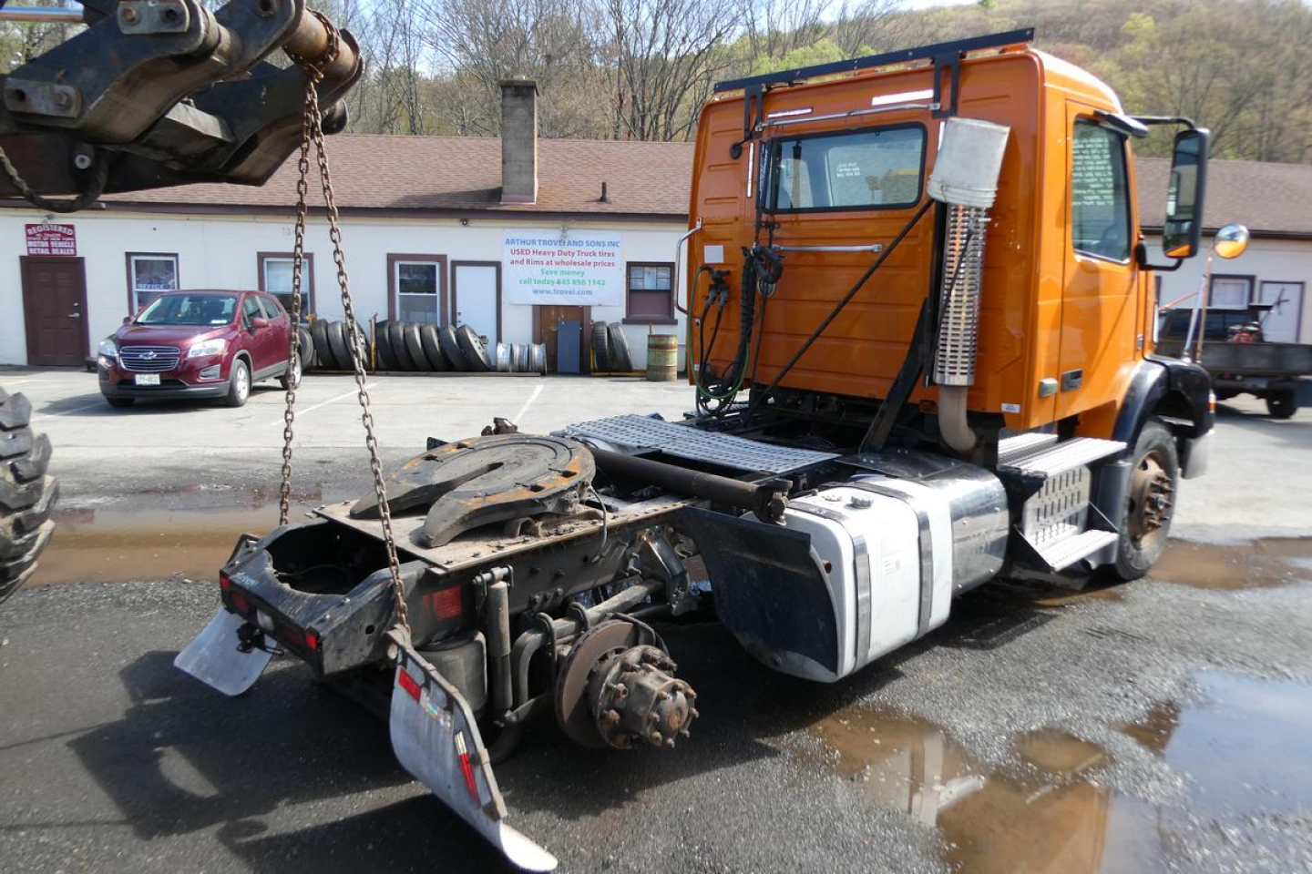 Orange Volvo VNM42T truck cab with exposed rear drivetrain and crane/arm visible, on a wet lot.