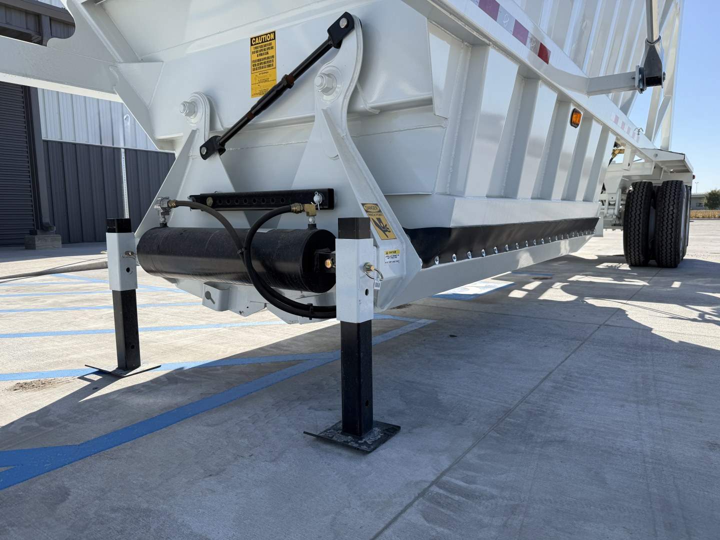 Trailer hitch view of Armor Lite SBD40 skeleton dump trailer showing hydraulic cylinder and support legs with yellow caution labels.