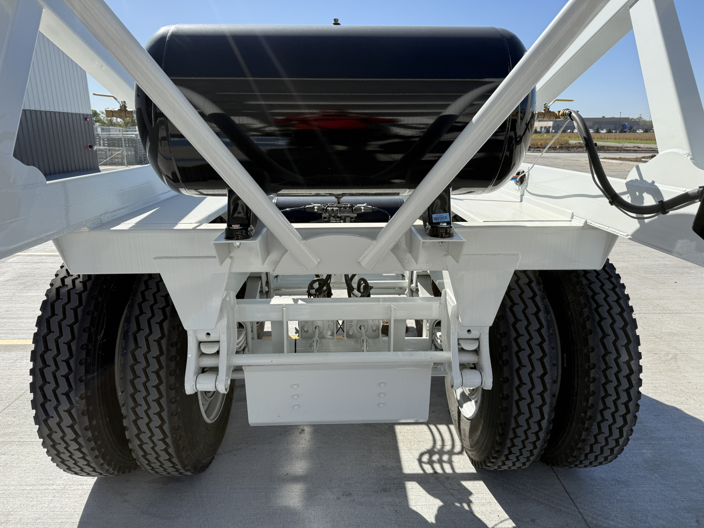 Dump trailer rear view showing dual tandem axles and suspension, black cylindrical tank, white frame, and rear hitch area.