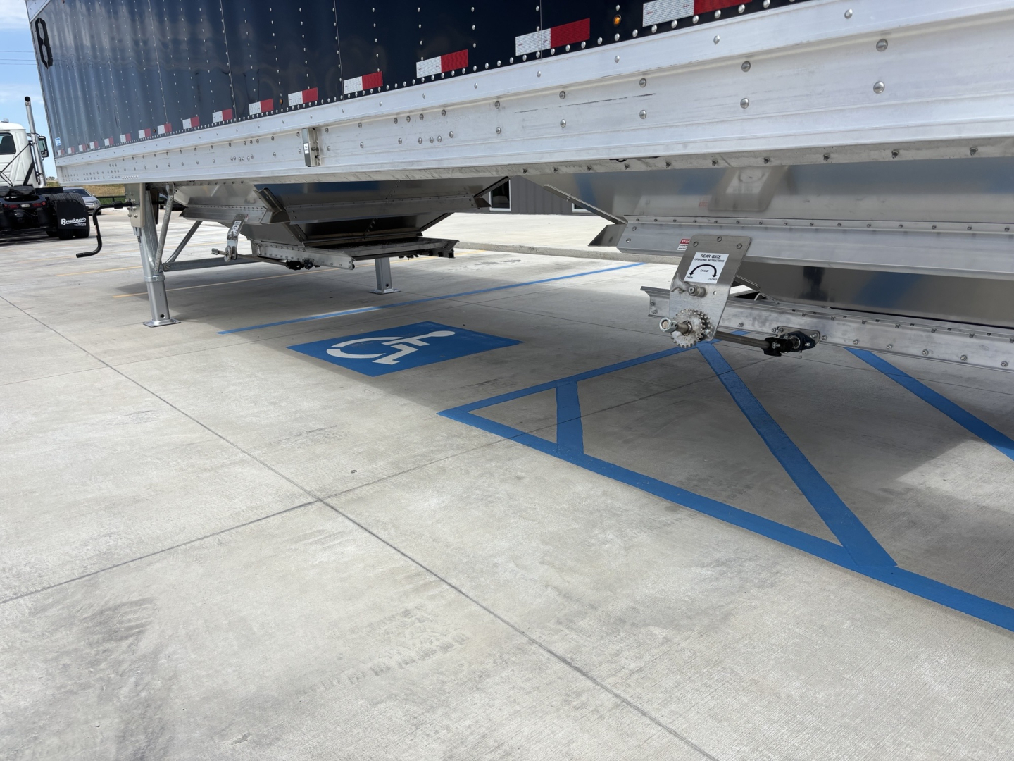 Truck trailer undercarriage and landing gear of a 42' aluminum grain hopper trailer with blue handicap parking marking visible on the concrete.