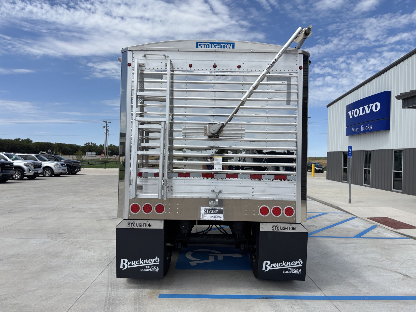 Trailer rear showing Stoughton hopper with aluminum grain trailer, 74-inch sides, vertical ladders and rear gate, license plate area, Bruckners logo mudflaps, and Volvo dealership in background.