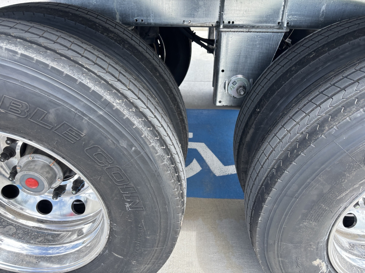 Dual rear axles of a trailer with large tires; some chrome rims; blue background with white logo; metal frame and suspension components visible.