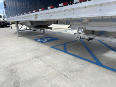 Truck trailer undercarriage and landing gear of a 42' aluminum grain hopper trailer with blue handicap parking marking visible on the concrete.