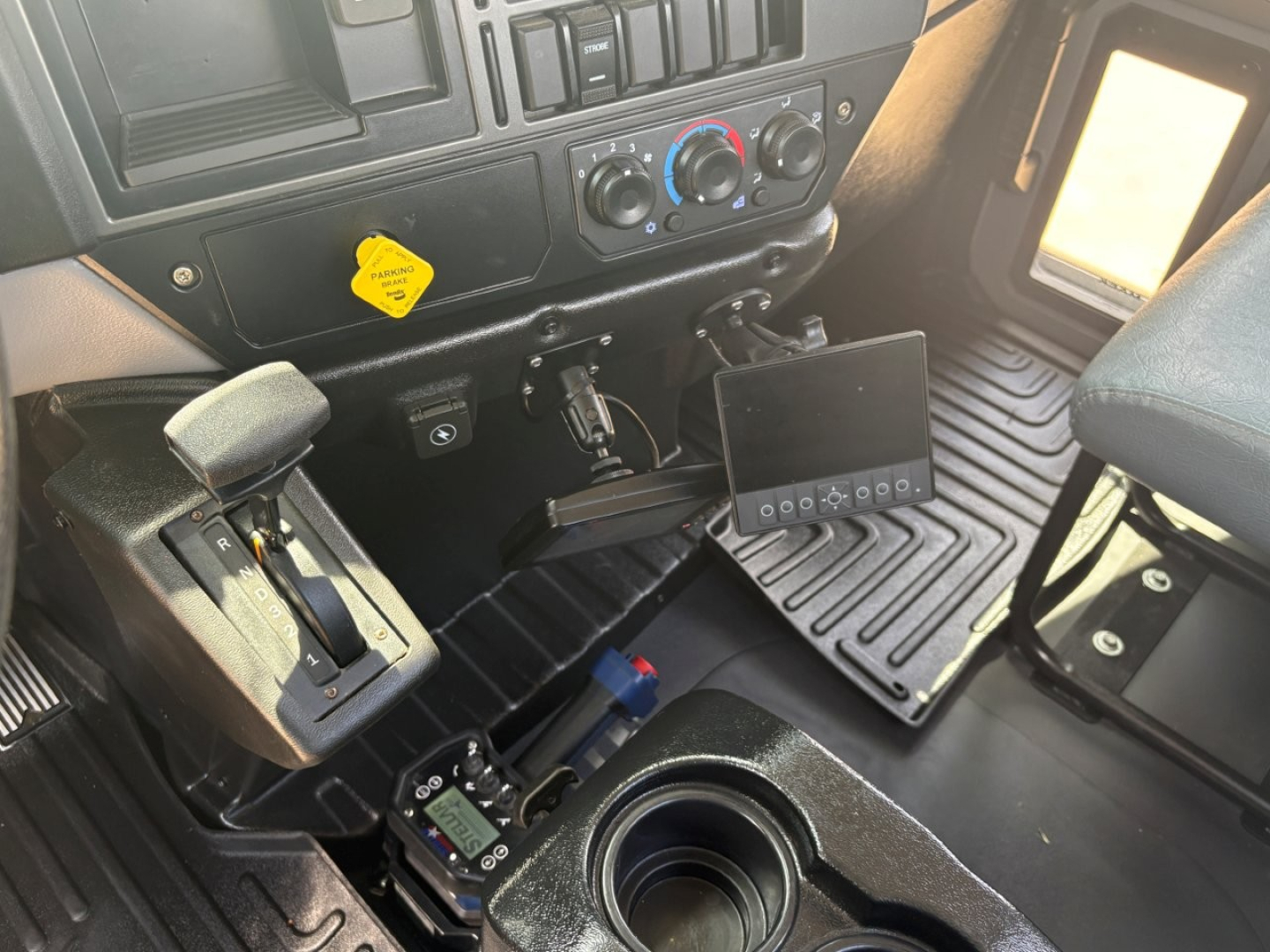 Interior of a 2024 Mack MD6 cab showing driver's seat area, steering column, climate controls, yellow warning sticker, and an aftermarket tablet/monitor mounted near the center console.