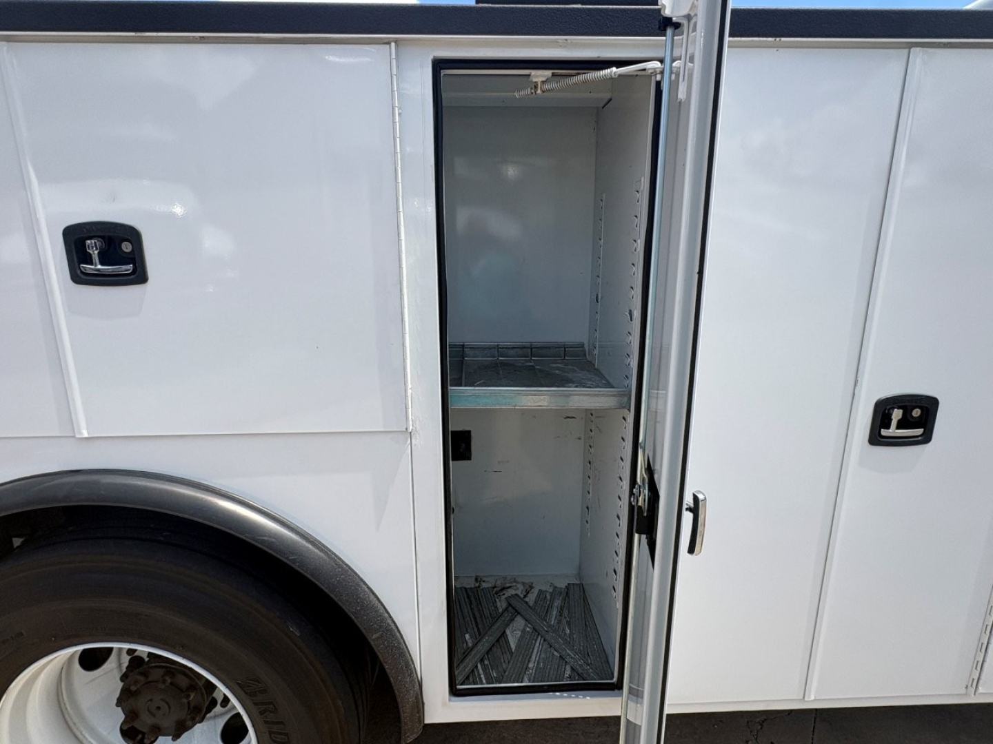 Service/utility truck side cargo box door open showing empty shelf inside cargo area of a white 2024 Mack MD6, with wheel well and aluminum interior flooring visible.