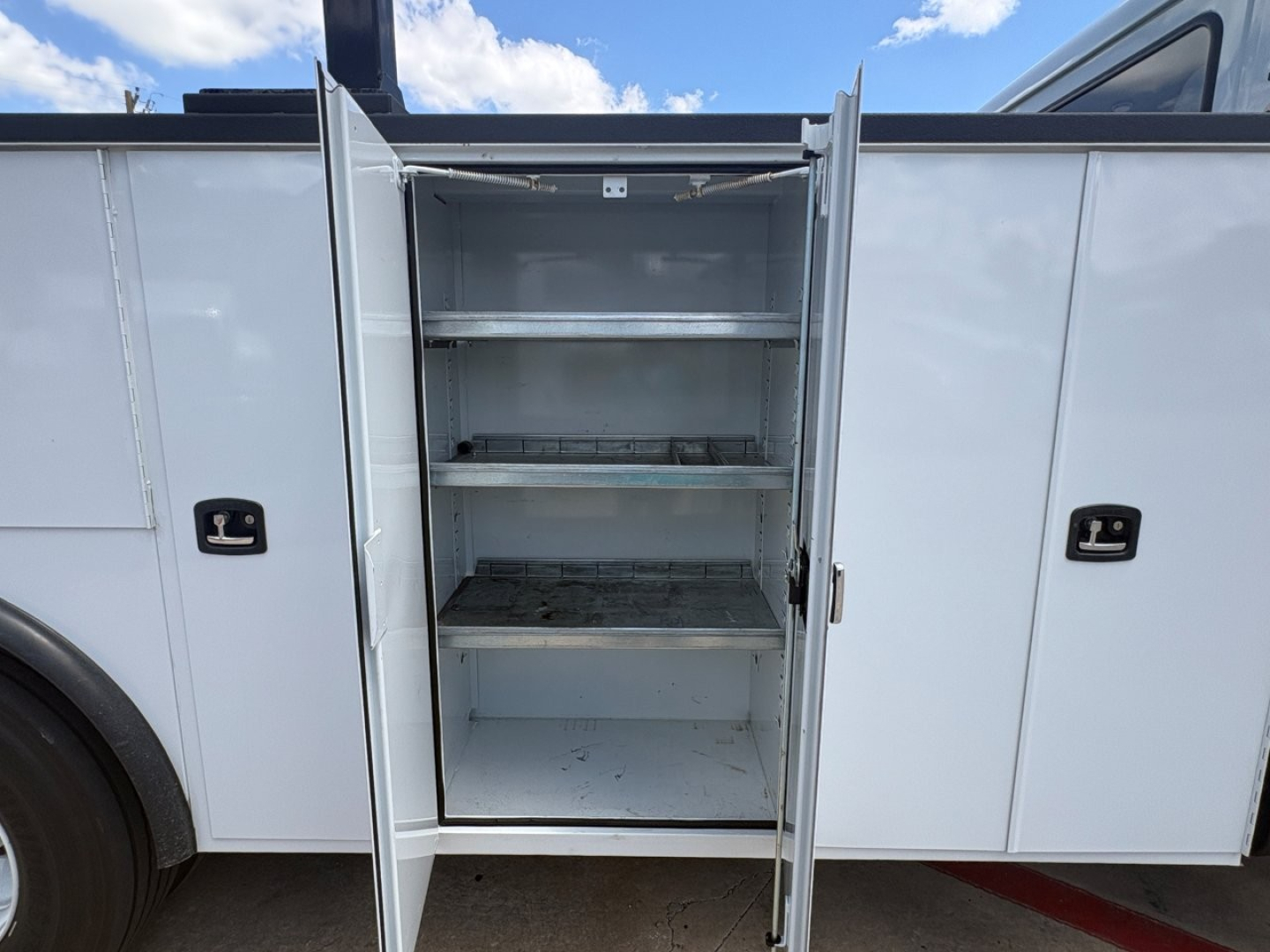 Service/utility truck body with an open side cabinet featuring multiple metal shelves and an empty top shelf.