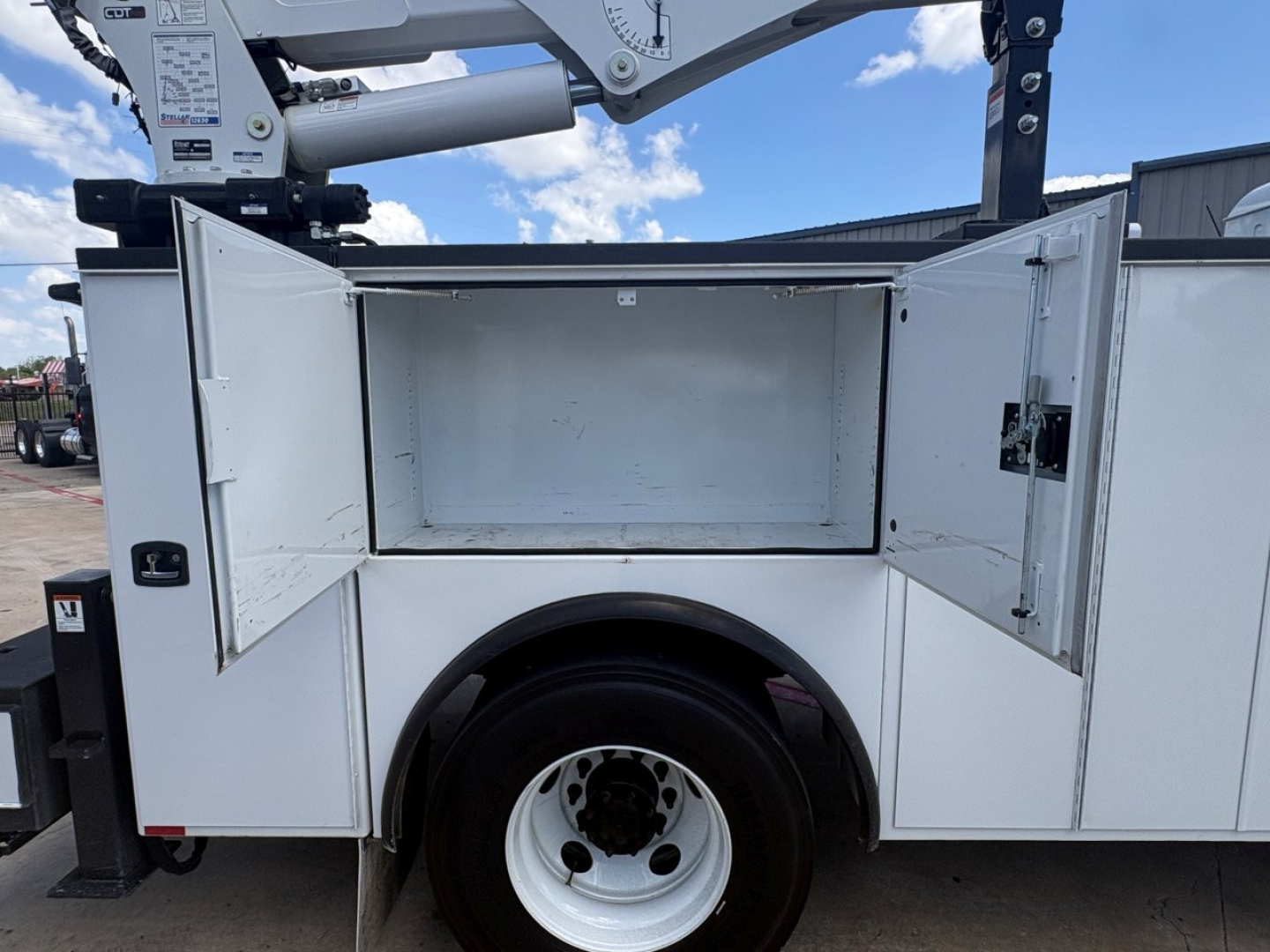 Mack MD6 service body truck side compartments open showing empty storage bay and hydraulic lifting mechanism above.