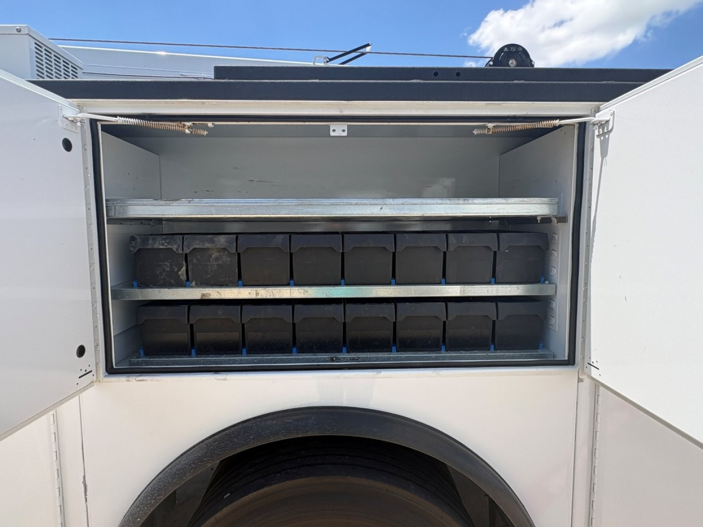 Truck utility body with open side storage compartments containing labeled bins and boxes in two shelves, Mack MD6 service vehicle, front wheel visible.