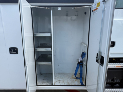 Service body of a Mack MD6 truck with open rear compartment showing shelves and a cargo area; blue strap and hooks inside, interior metal walls, shelves on left side.