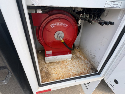 Backyard view of a red Reelcraft hose reel mounted inside a service truck compartment, with hoses and metal fittings visible; floor shows rust and dirt.