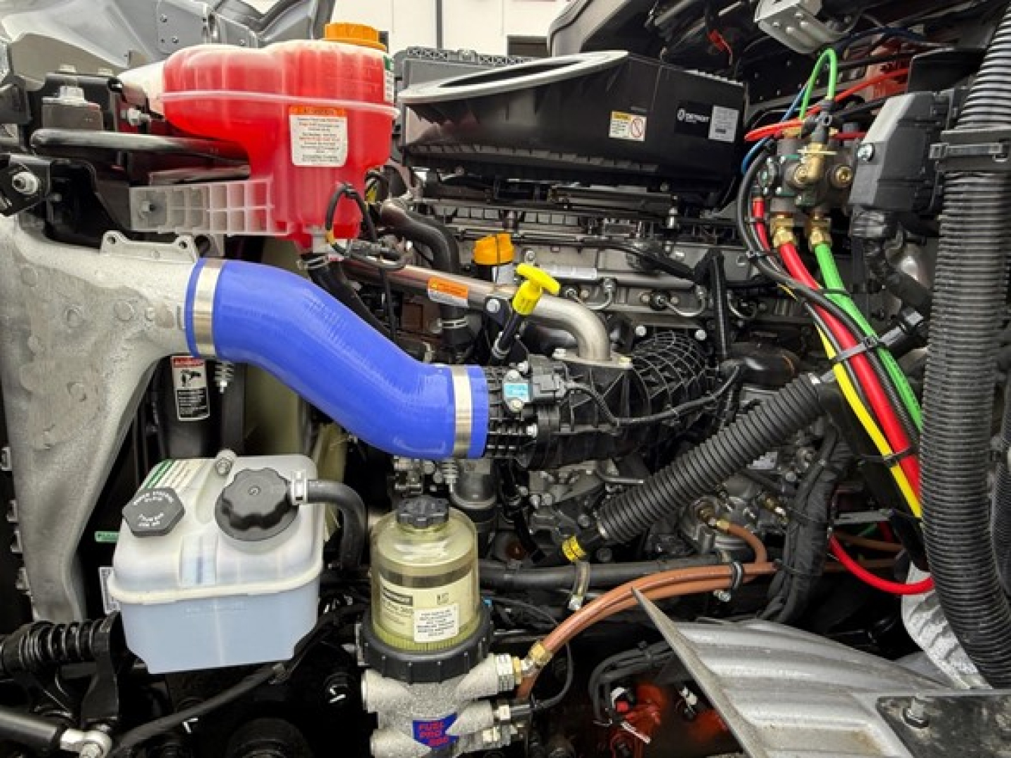 Engine bay of a Western Star 47X dump truck showing blue intercooler/duct hose, red coolant reservoir, white coolant overflow bottle, various hoses, wiring, and a yellow dipstick, with a gold/green coolant reservoir cap.