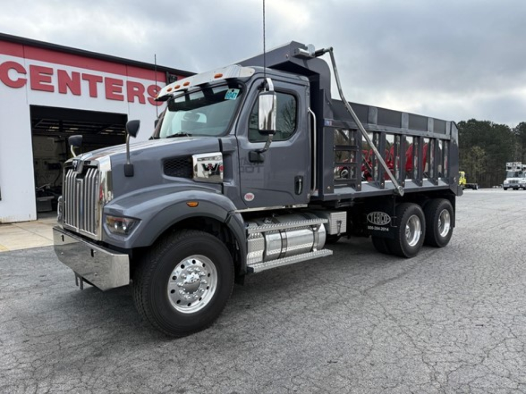 Dump truck in showroom lot showing 2026 Western Star 47X with gray cab and dump bed, chrome exhaust stack, dual rear axles, front chrome bumper, large mirrors.