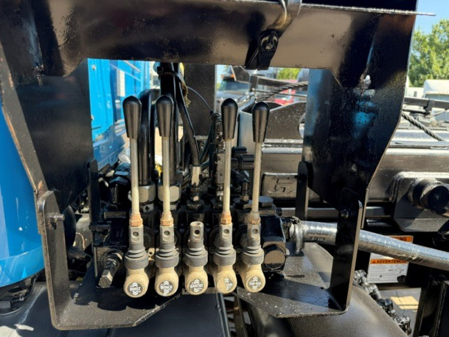 P5700 Mack GR64B control levers with labels and fittings in a hydraulic valve bank under a roll-off truck body.
