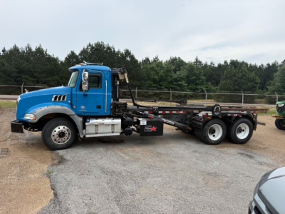 Blue Mack GR64B roll-off truck with crane arm and flatbed chassis, side view showing cab, fuel tanks, and dual rear axles.