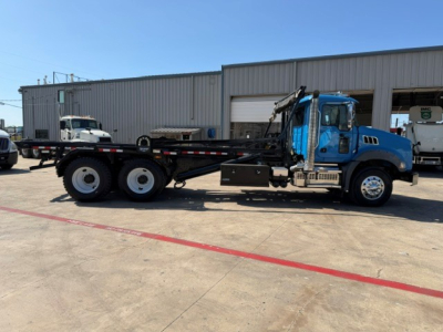 Truck side view showing a blue 2021 Mack GR64B roll-off wrecker chassis with six wheels, crane, and flatbed at a service yard.