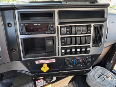 Dashboard panel of a 2026 Mack MD7 truck showing the audio head unit, multiple switches, climate controls, and a storage compartment.