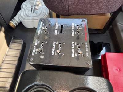 Control panel with multiple toggle switches labeled for front left, front right, rear left, and rear right directions, mounted in a metal box on a truck interior floor.