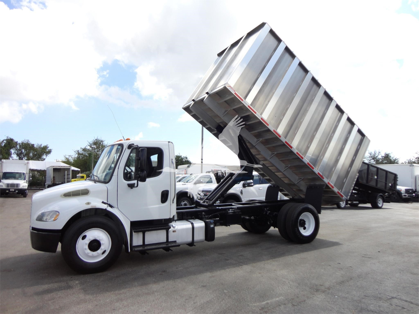 Freightliner M2 106 medium-duty truck with tilted dump bed in a yard showing chassis and cab, multiple heavier vehicles in background.