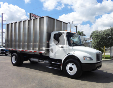 Freightliner M2 106 chassis with large metal dump bed, white cab, side view showing logo on door and cargo bed; truck on lot under blue sky.