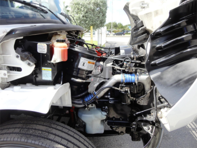 Freightliner M2 106 engine bay view showing coolant reservoir, battery, hoses, and engine components with open hood, partial view of chassis.