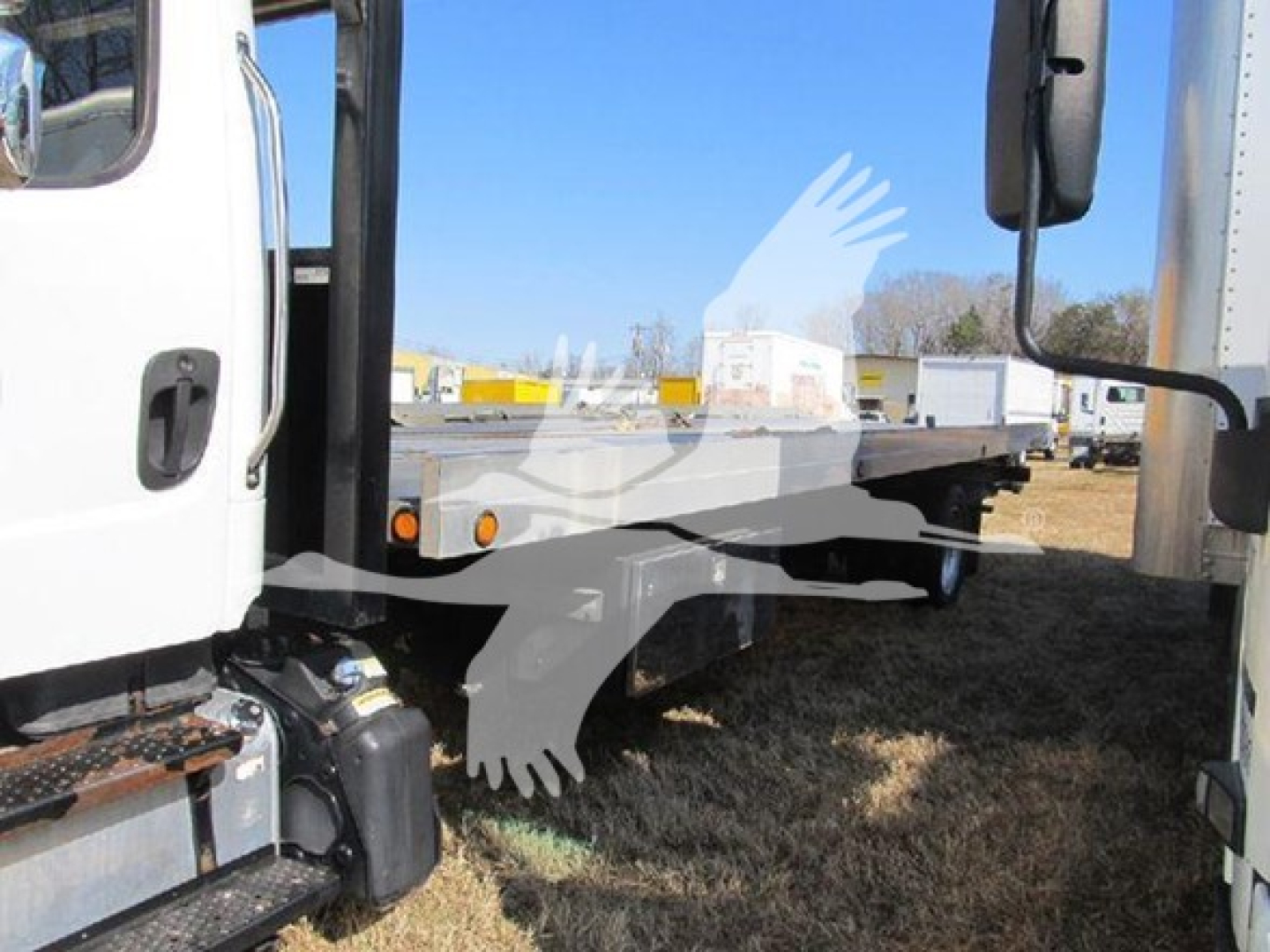 Flatbed auto carrier truck with white cab and gray flatbed, showing side mirror and lower steps. Windowed door on left; graphic watermark of an eagle overlay. Clear blue sky.