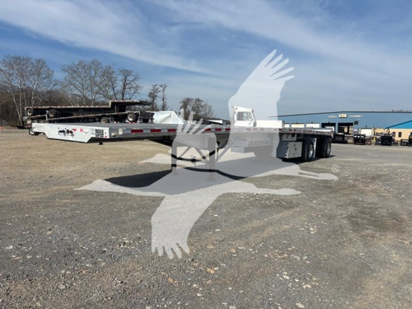 Flatbed trailer in a yard, side view, 3-axle, low-profile tandem with silver rails and white deck; VIN and stock info not visible; no brand text legible.