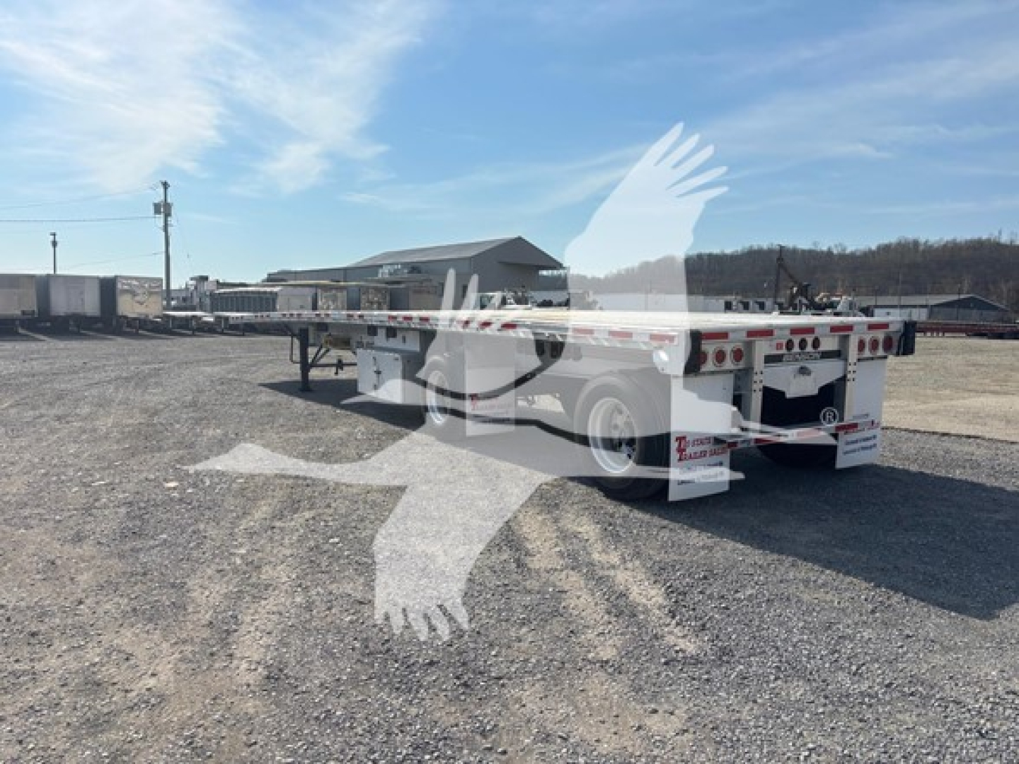 Low-angle view of a Benson flatbed trailer in a gravel lot, showing rear and trailer frame with wheels, side rails, and rear lights.