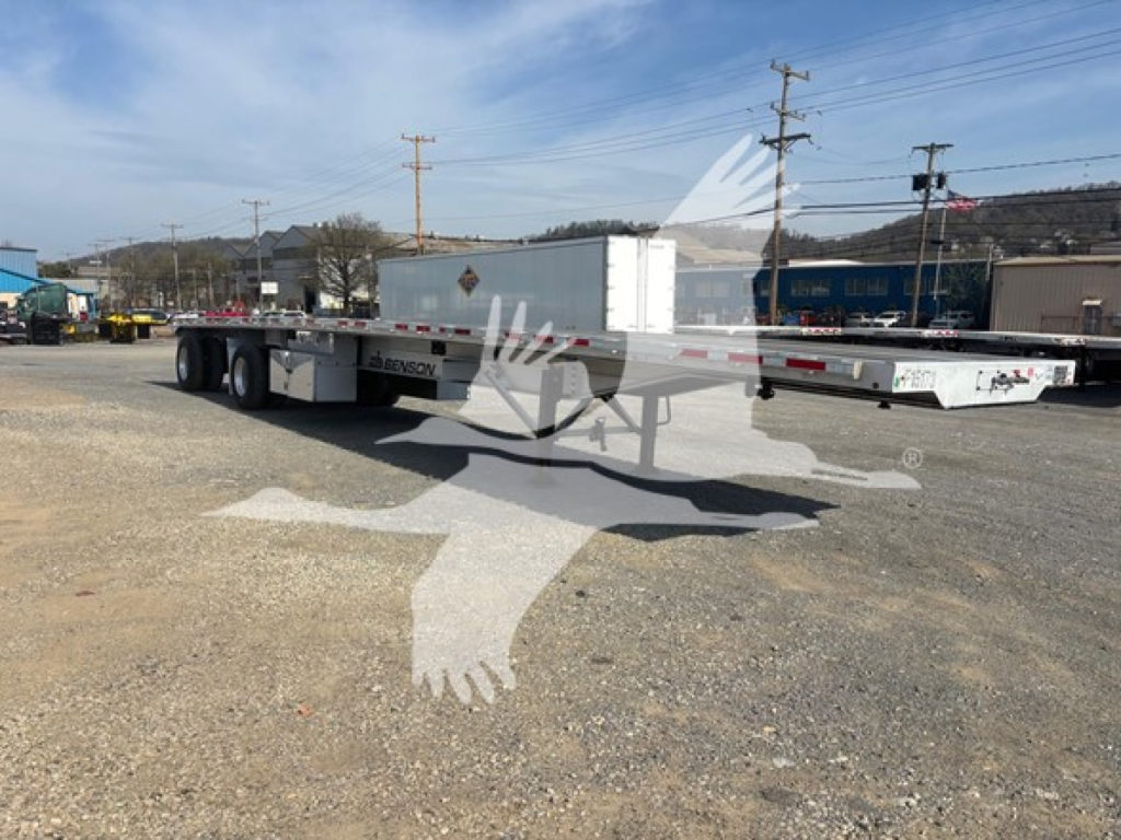 Flatbed trailer in a gravel lot showing Benson brand with tandem axles and a white toolbox mounted on the front.