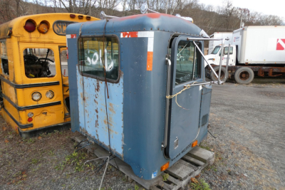 Blue truck cab shell with rear window and side window, rust patches, tied roof, sitting on pallets in a yard beside a yellow bus and other vehicles.