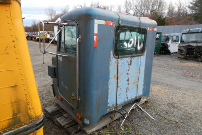Cabin-style truck sleeper unit blue with rectangular windows and rust patches, viewed from rear left; attached hoses on ground, other trucks in background.