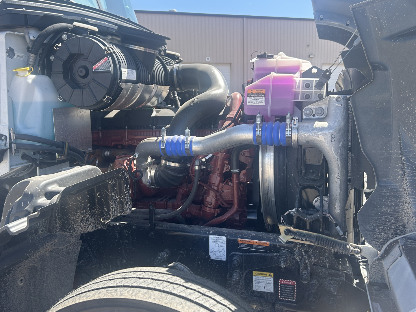 Mack GR64FT engine bay showing turbocharger, intake hoses, coolant reservoir, purple coolant or DEF reservoir, and engine block with orange components.