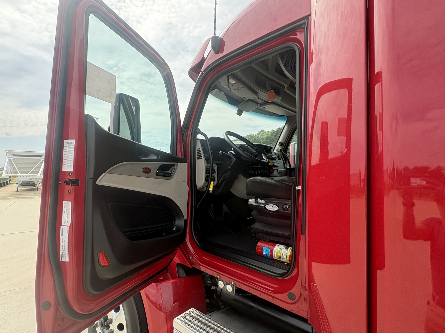 Truck cab doorway of a red 2023 Peterbilt 579 with open door showing steering wheel and dashboard; interior items visible near floor.