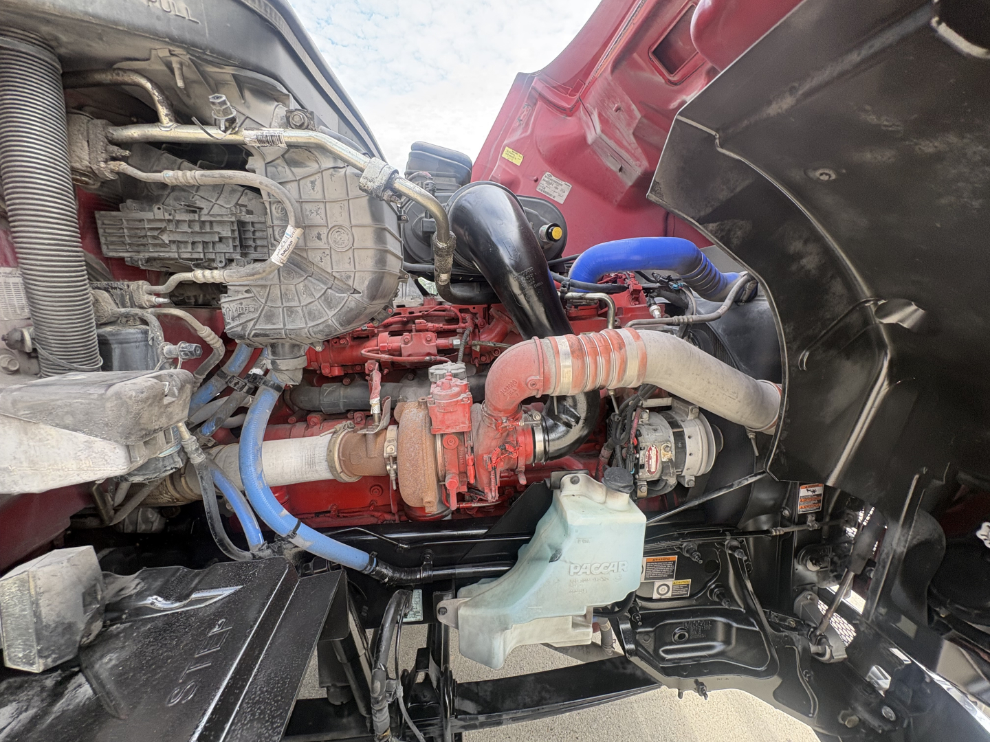 Peterbilt 579 engine bay showing diesel engine, blue hoses, red intake, coolant reservoir, and surrounding components.