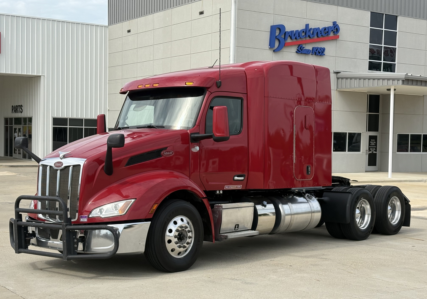 Peterbilt 579 tractor in red in front of truck dealership building. Front bumper guard, large chrome grille, dual fuel tanks, and three rear axles visible.