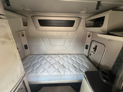 Truck sleeper cab interior of a 2023 Peterbilt 579 showing a bed with light-gray quilted mattress and side storage compartments.