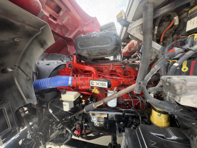 Peterbilt 579 engine bay showing red X15 diesel engine with blue hose, various hoses, wiring, and components visible.