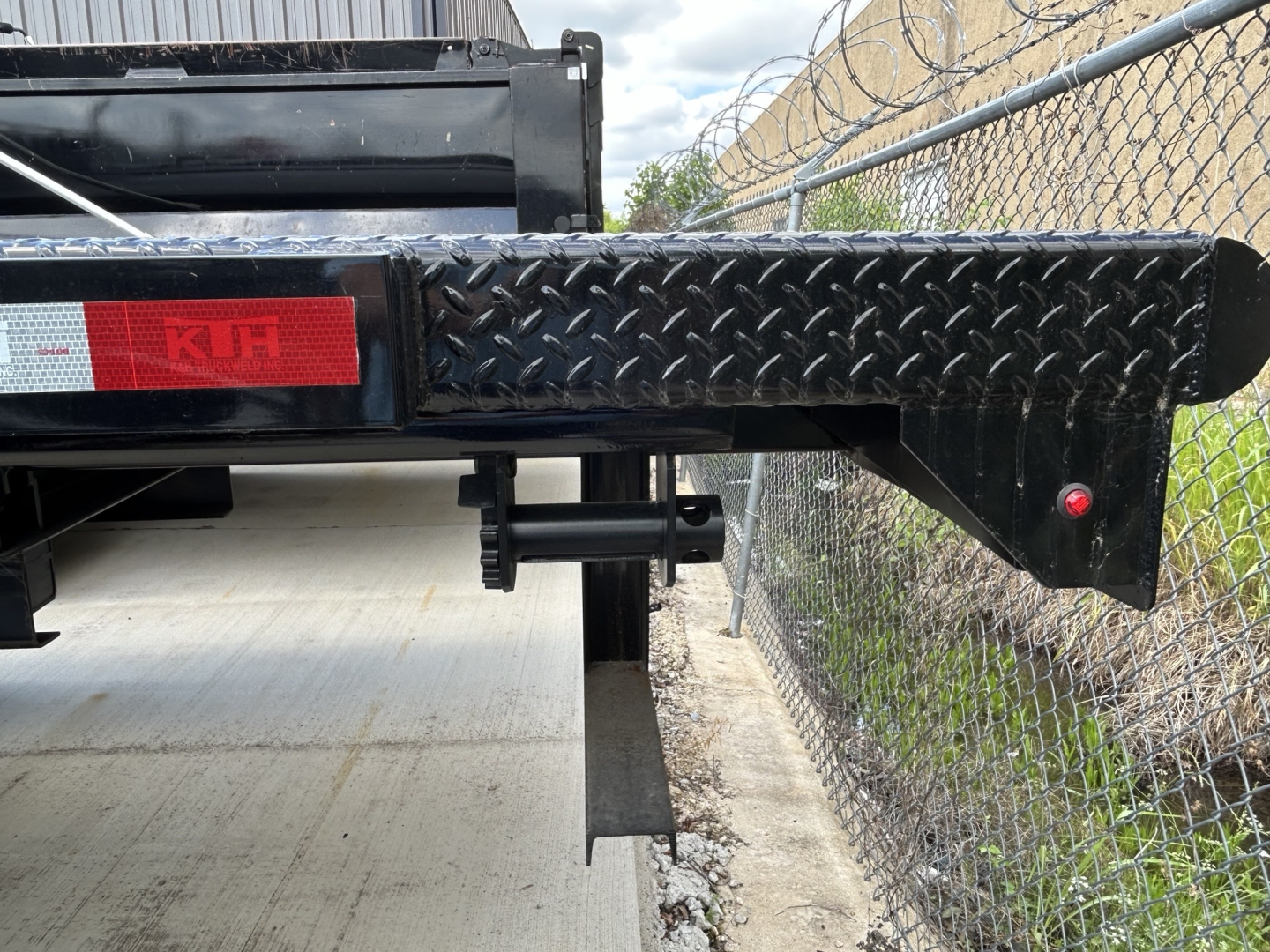 Flatbed trailer rear-left side of a 2026 Mack MD6 showing diamond plate steel step and extendable hitch with red reflector and K&N badge.