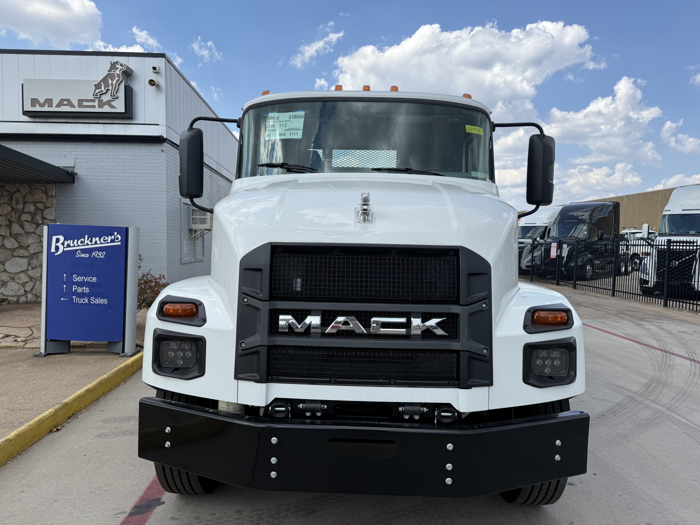 Front view of a white 2026 Mack MD6 truck showing bold black grille with MACK lettering, large black bumper, side mirrors, and factory sticker on windshield.